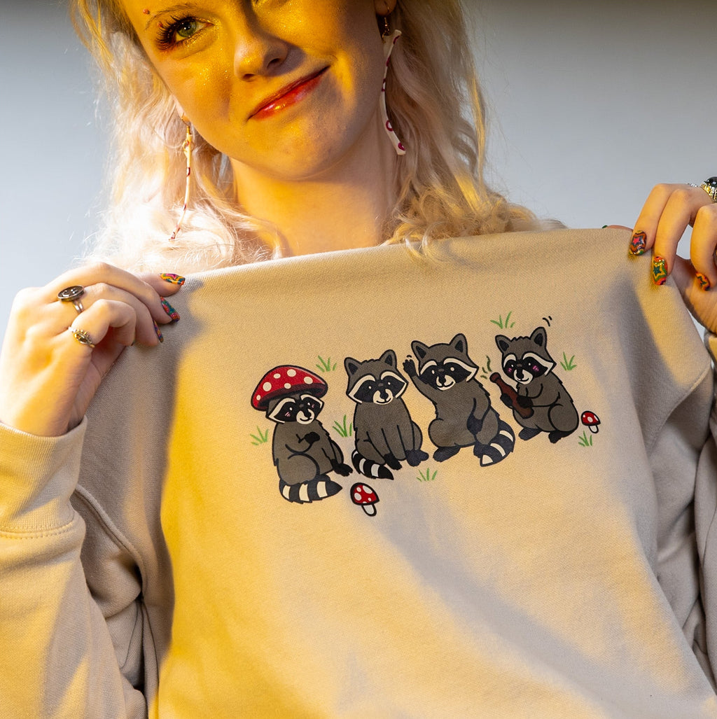 Person holding a light-colored sweatshirt with raccoon and mushroom design.