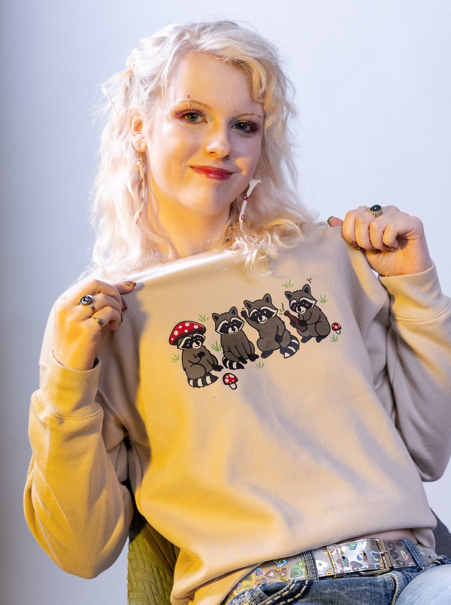 Woman holding a beige sweatshirt with raccoon and mushroom design against a light gray background