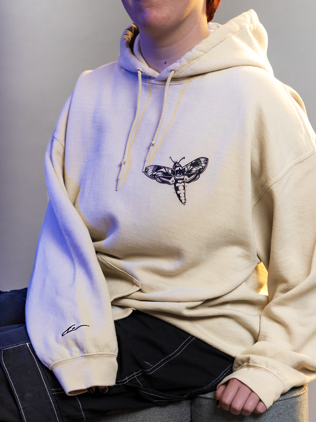 Person wearing a beige hoodie with a black moth design on a gray background