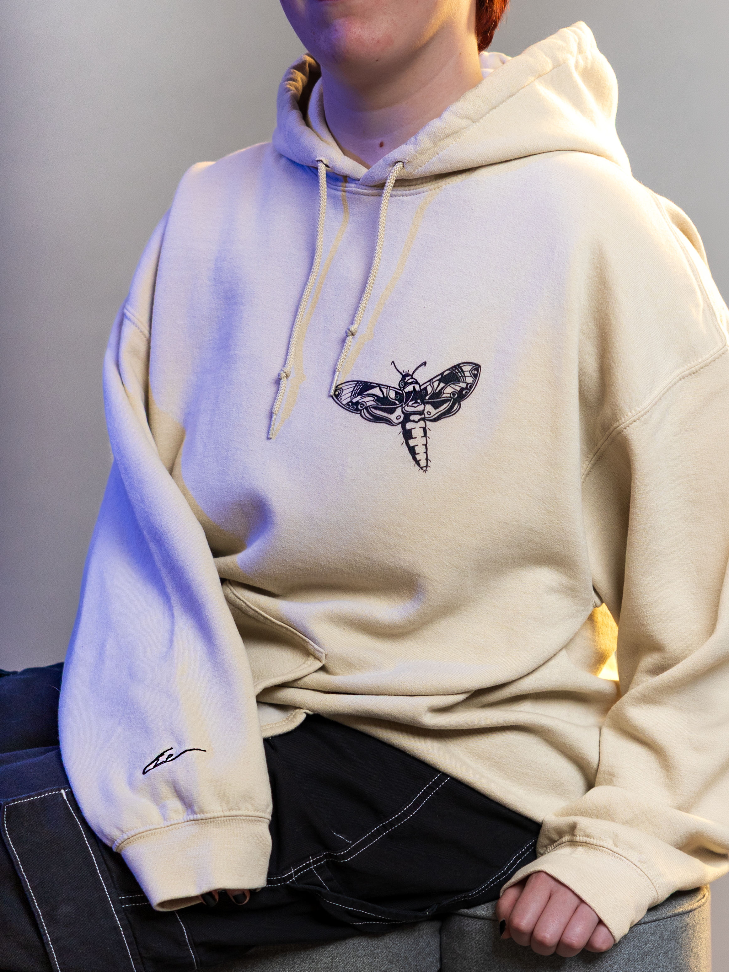 Person wearing a beige hoodie with a black moth design on a gray background