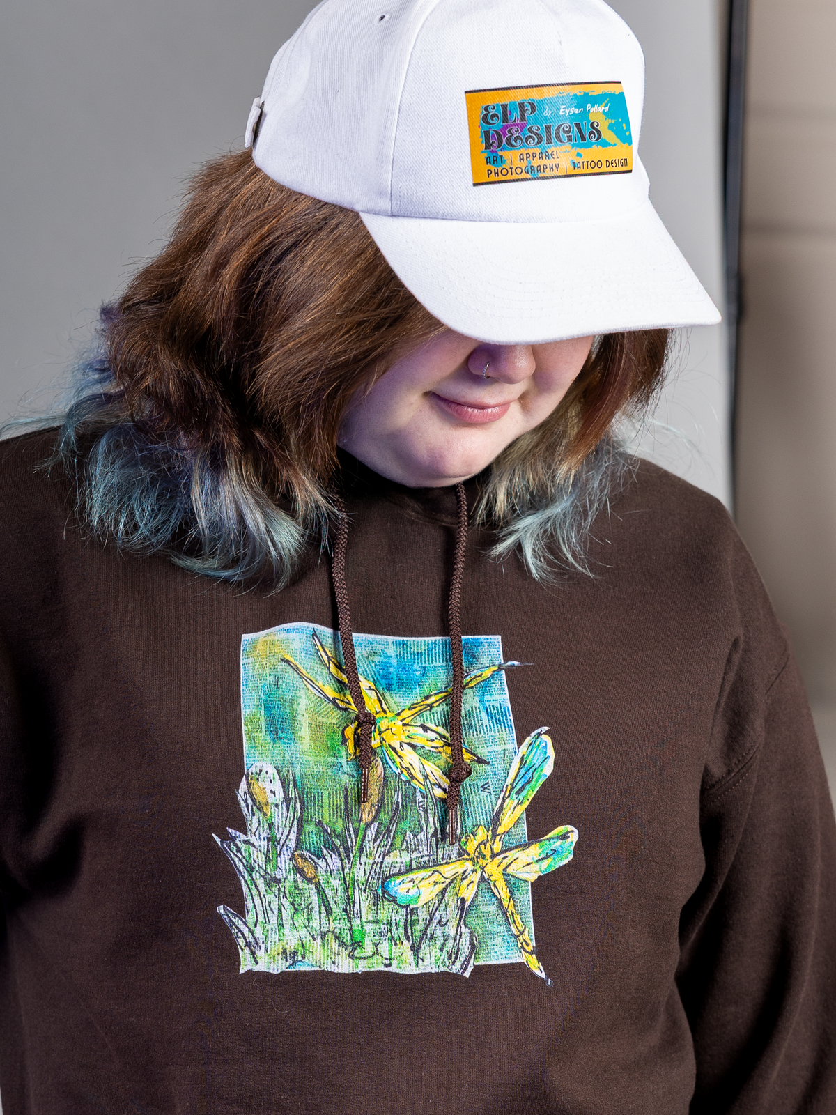 Person wearing a brown hoodie with a dragonfly design and a white cap with a logo.