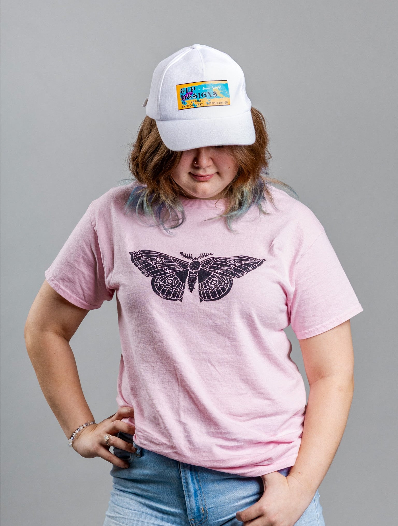 Person wearing a pink t-shirt with a textured black moth design and a white cap with a colorful logo on a gray background